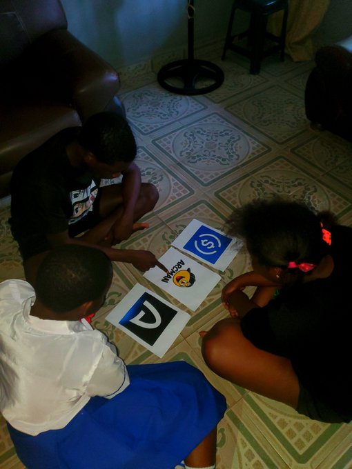 Three children sit on a tiled floor looking at printed cards displaying letters 'd', a battery graphic with the text 'Recharg' and 'e', suggesting an educational activity.