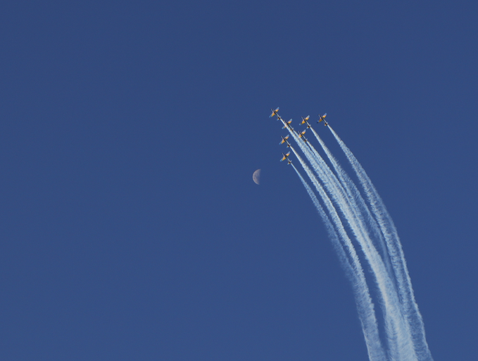 A photograph of six jets in formation trailing white smoke across a deep blue sky, with the moon visible to their left.