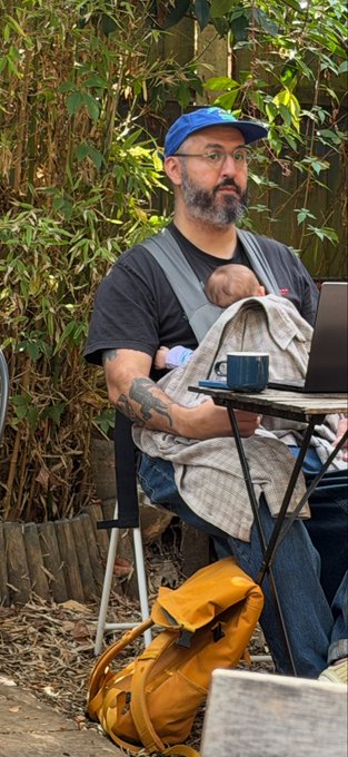 A man with a beard, glasses, and blue cap sits in a chair wearing a baby carrier, with a yellow backpack at his feet and a laptop and coffee mug on a small table.