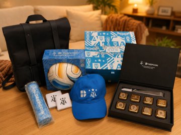 A collection of promotional merchandise including a black backpack, a blue football, a blue cap, a water bottle, keychains, and a boxed set of chocolates arranged on a wooden table.