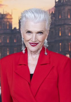 A stylish older woman with short white hair poses confidently in a bright red blazer and statement earrings, with a sunset-lit cityscape behind her. The image corresponds to the post referencing Maye Musk, though no identifying text or name is visible on the image itself.
