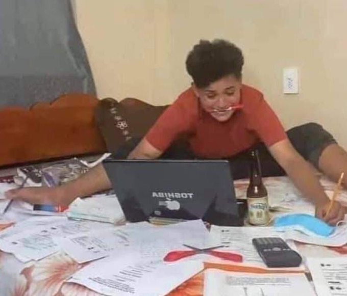 The image shows a young man sitting on his bed with a laptop open in front of him. He appears to be working or studying, surrounded by various items such as papers, books, and a bottle. The scene suggests that he might be multitasking between different tasks or projects while using the laptop for research or communication purposes.