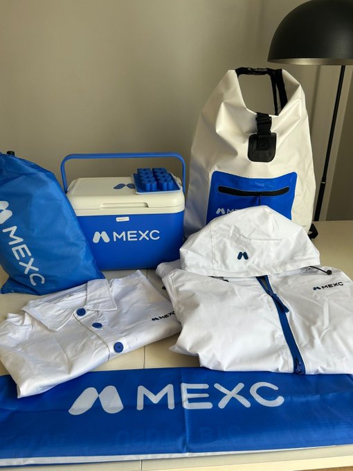 A collection of MEXC-branded merchandise, including a blue cooler, dry bag, cap, jacket, polo shirt, and banner, is displayed on a table alongside a black lamp, showcasing the promotional items received in the package.