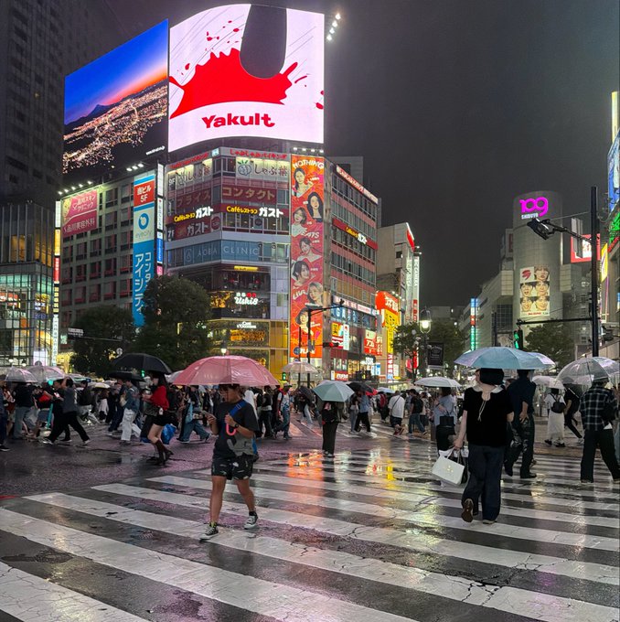 A nighttime view of the famous Shibuya Crossing in Tokyo, Japan, featuring the iconic Yakult advertisement and other illuminated billboards as crowds of pedestrians with umbrellas cross the street in the rain.