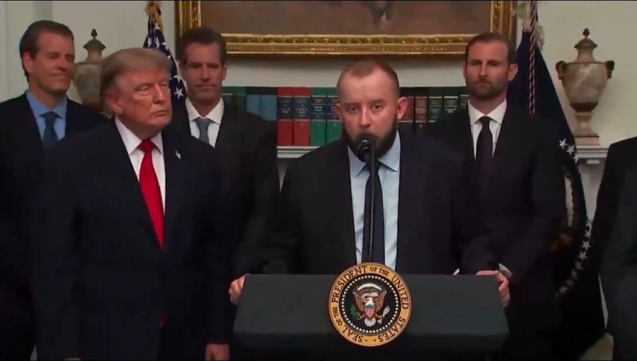 A man speaks at a podium bearing the Seal of the President of the United States, with President Donald Trump standing to his left and Secret Service agents behind them.