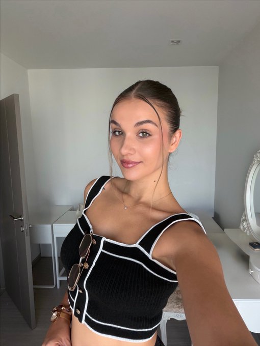 A young woman takes a mirror selfie in a bedroom, wearing a black and white striped knit crop top and matching skirt with buckle details, accessorized with layered necklaces.