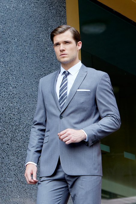 A man stands outdoors wearing a tailored gray suit with a white shirt and a blue-and-gray striped tie.