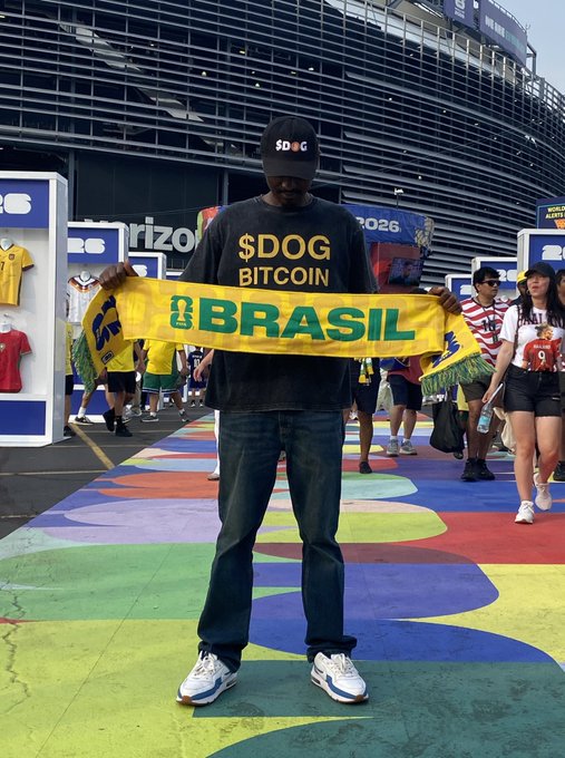 A man wearing a "$DOG BITCOIN" shirt and a "$DOG" cap holds up a green and yellow "BRASIL" scarf at what appears to be a World Cup 2026 venue, embodying the post's "DOG•GO•TO•THE•MOON" rallying cry with his full DOGE-branded attire at the global sporting event.