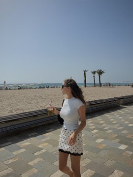 A woman in a white top and patterned skirt holding a drink, walking along a beachside promenade with palm trees and the ocean in the background.