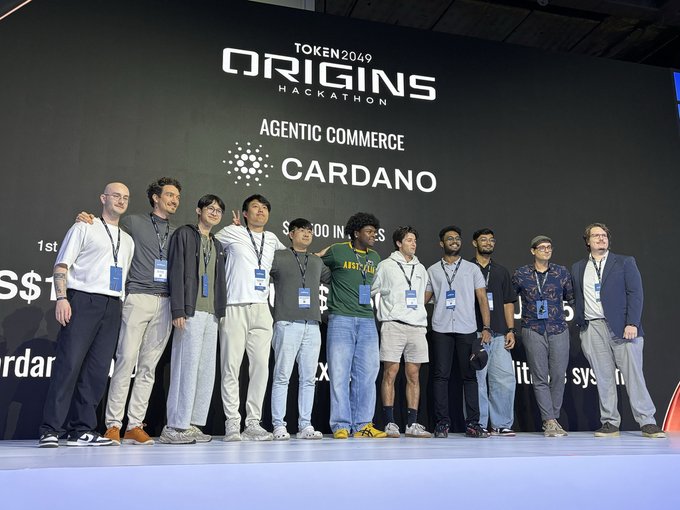 A diverse group of approximately 13 developers pose together on stage at the TOKEN2049 Origins Hackathon, with the backdrop displaying "AGENTIC COMMERCE CARDANO." The team's celebratory stance reflects the post's announcement about the Cardano track's strong participation, with 2 of the top 5 projects emerging from this group of builders who chose Cardano from five available ecosystems.