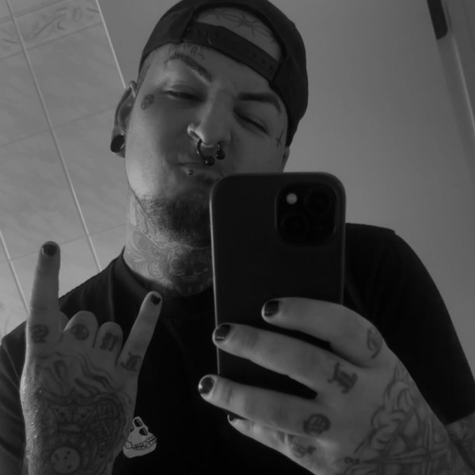 A black and white mirror selfie shows a tattooed man wearing a backwards cap, holding up a middle finger while taking a photo with his phone.
