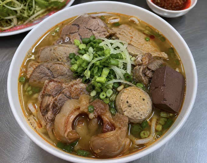 A close-up photo of a Vietnamese noodle soup bowl containing beef, pork, meatballs, blood cubes, and green onions in broth, with side dishes of vegetables and chili sauce.