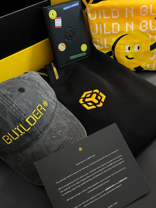 The image features a black box with yellow text that says "Build-n-Build" and includes a hat and a sweatshirt inside. The hat appears to be made by Builder, while the sweatshirt is yellow and has a logo on it. A letter is also present in the scene, which seems to be part of an email or message related to the merchandise.