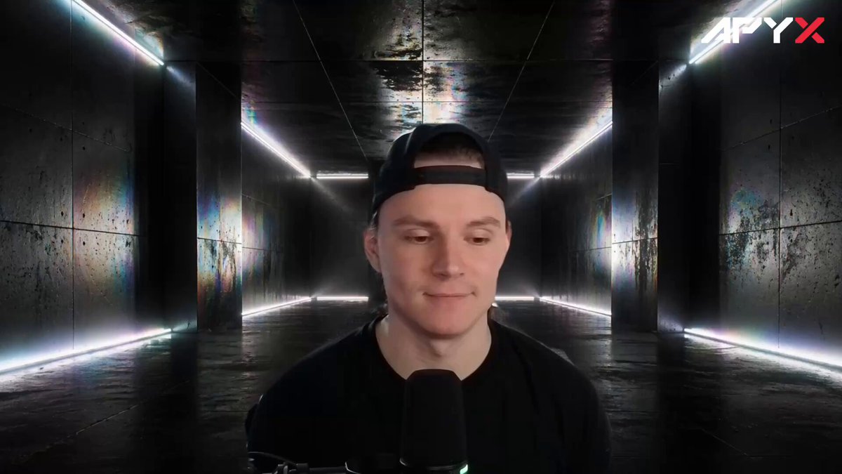 The image features an adult male wearing a black hat and shirt, standing in front of a microphone with his face lit up by a spotlight. The backdrop consists of a series of dark hallways lined with bright lights that create an eerily atmospheric setting. The man appears to be speaking into the microphone or participating in a live stream event.