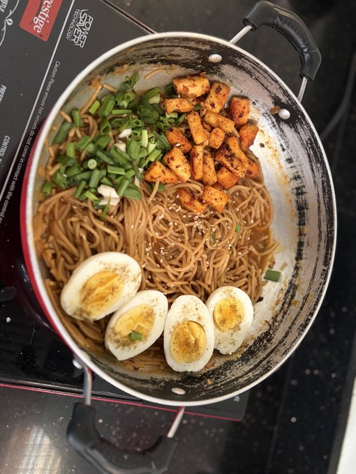 Top-down view of a pot containing noodles topped with tofu cubes, chopped green onions, sesame seeds, and halved boiled eggs.