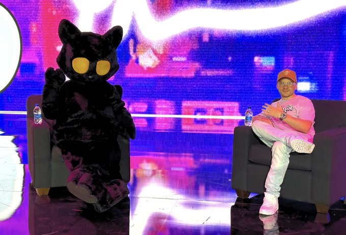 Two figures sit in armchairs on a neon-lit stage: a person in a black furry mascot costume with glowing yellow eyes and a man wearing a cap, glasses, and white outfit, both with water bottles nearby.