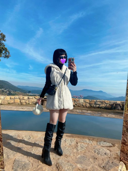 A person with short black hair poses for a mirror selfie at a scenic overlook, wearing a white fuzzy vest, black knee-high boots, sunglasses, and a black face mask, while holding a phone with a pink heart emoji covering their face—likely a reintroduction post aligning with the tech/crypto-themed profile of a retired developer supporting Monad and $MON.