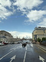 A street view photo taken from inside a vehicle, showing a wet road with directional arrows, flanked by classical buildings and a partly cloudy sky in the distance.