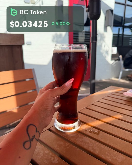 A hand with pink nails holds a tall glass of dark beer on a wooden patio table, with an overlay showing 'BC Token' at '$0.03425' up 5.00%.