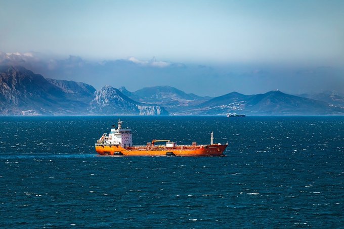 An orange cargo ship sails on blue ocean waters with a rugged mountain range visible in the misty background.
