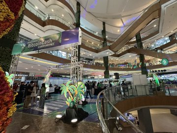 An interior view of a multi-story shopping mall featuring a 'Bitcoincash' banner hanging from the ceiling, with people gathered around a display in an atrium filled with tropical plants.