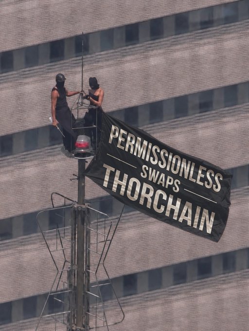 Two activists scale a communications tower outside a high-rise building, unfurling a banner reading "PERMISSIONLESS SWAPS THORCHAIN" in protest-style lettering. The image aligns with the "GM" post by showcasing a bold, attention-grabbing crypto-related demonstration promoting the THORChain decentralized exchange.
