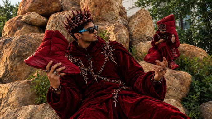 A person wearing an elaborate red velvet robe, spiked crown, dark sunglasses, and spiked jewelry reclines on rocks while holding a matching red pillow, with another similarly dressed figure seated on rocks in the background.