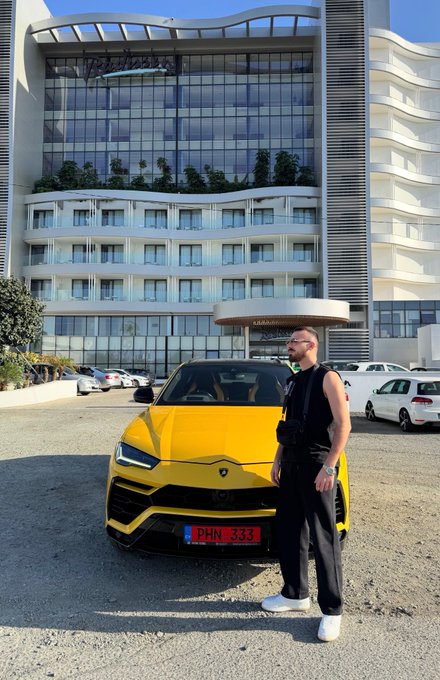 A man in a black sleeveless outfit stands proudly beside a bright yellow Lamborghini Urus with the license plate "PHN-333," in front of a modern glass-facade hotel labeled "KARAVAN." The luxury supercar and upscale setting reinforce a confident "Gm friends" greeting, showcasing a lifestyle of wealth and style.