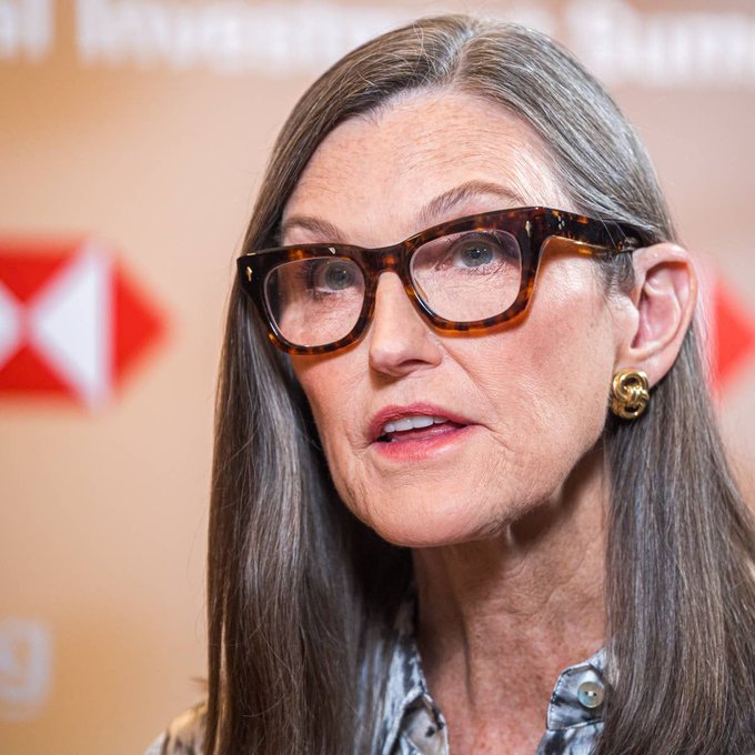 Cathie Wood, known for her bullish stance on Bitcoin and founder of ARK Invest, is shown wearing tortoiseshell glasses and gold earrings, appearing to speak at what looks like a press event with HSBC branding in the background. The image accompanies an article about her optimistic response to Bitcoin "dead cat" warnings, reinforcing her reputation as a prominent advocate for the cryptocurrency's long-term potential.