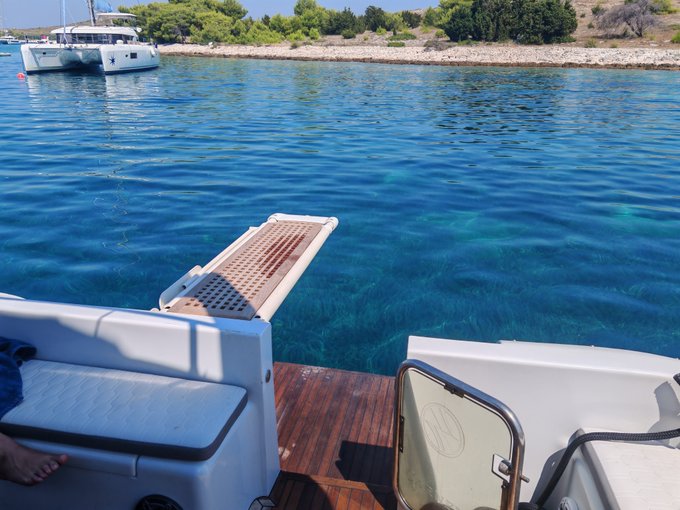 A view from a boat shows a wooden gangplank extending toward clear turquoise water, with a catamaran anchored near a tree-lined shore in the background.