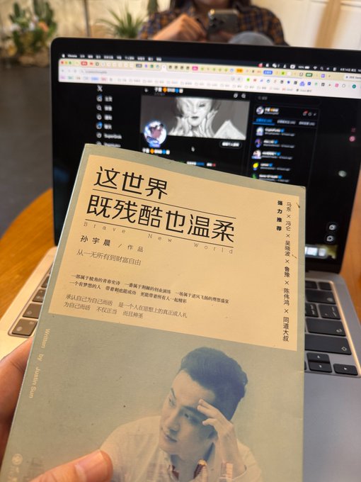 A hand holding a Chinese book titled '这世界既残酷也温柔' (Brave New World) by 孙宇晨 (Justin Sun) in front of an open laptop displaying a webpage.