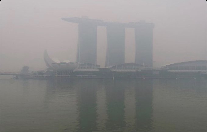 A hazy, fog-covered view of the Marina Bay Sands hotel and the ArtScience Museum in Singapore, reflected in the calm water below.