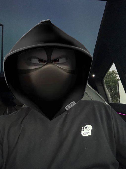 A person wearing a black hoodie with a cartoonish ninja-like mask design and a small skull logo on the chest, sitting in a Tesla at night.