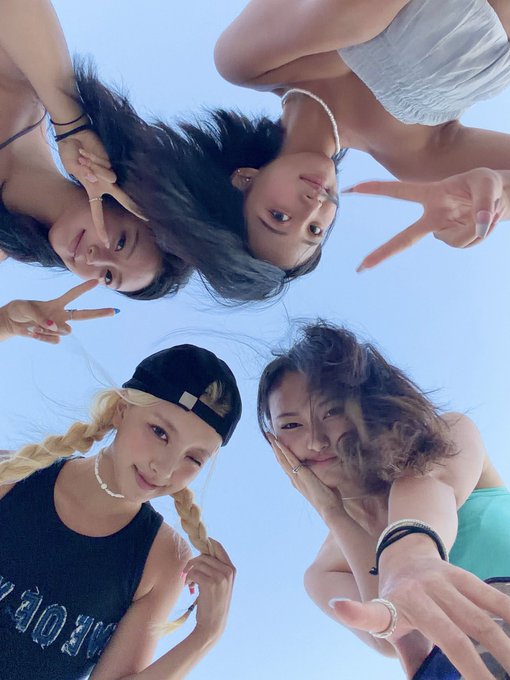 A worm's-eye view photo of four young women posing playfully against a clear blue sky, with one wearing a black cap and blonde braids.