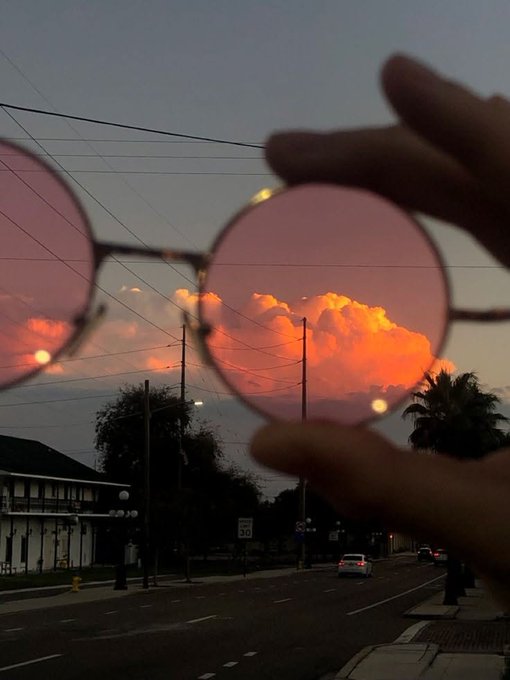A hand holds up pink-tinted round sunglasses against a stunning sunset with vibrant orange clouds over a city street at dusk.