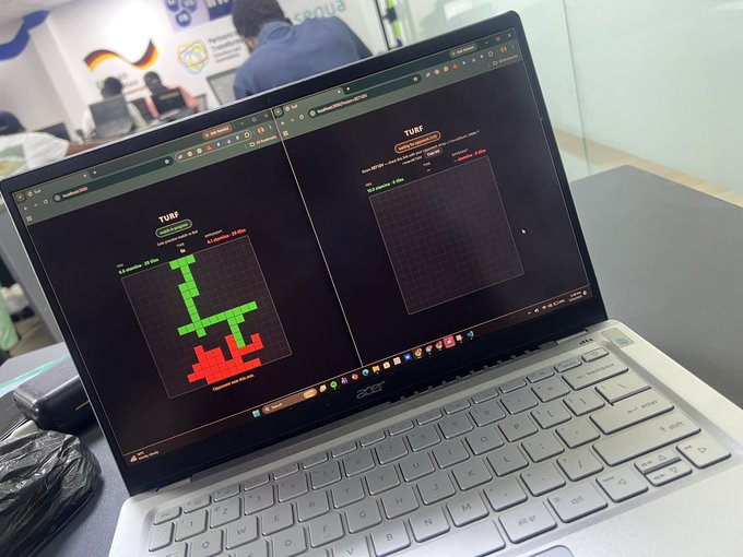 An Acer laptop displays two browser windows running "TURF" game prototypes, with the left screen showing a green and red blocky arena and the right showing an empty grid—capturing the milestone moment when the project moved from concept to a working demo.
