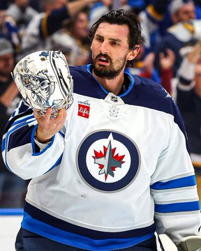 A bearded hockey player wearing a white Winnipeg Jets jersey holds up his goalie mask in celebration, with a Life-s sponsorship patch visible on his chest.