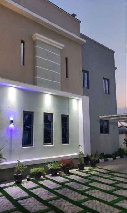 An exterior view of a two-story modern home captured at dusk, featuring large windows, illuminated accent lighting, and a patterned stone driveway lined with small potted greenery.