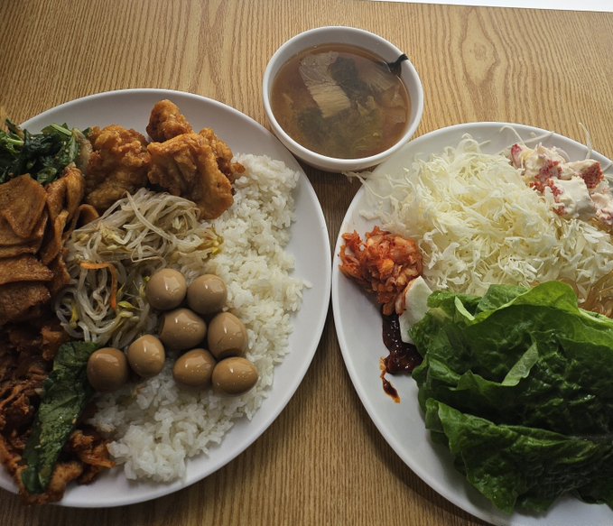 Two plates of food on a wooden table featuring rice, fried items, quail eggs, bean sprouts, kimchi, shredded cabbage, lettuce wraps, and a bowl of soup.