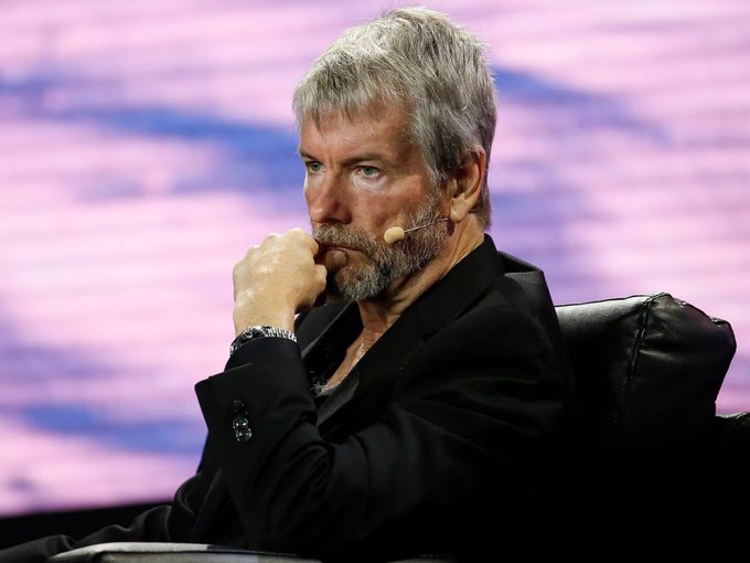 In this image, we see an older man sitting in a chair with his hands tucked into his pockets. He appears to be deep in thought or concentration. The background of the picture features pinkish-purple hues, which may indicate that he is looking at something on a screen behind him. The text in the photo reads "ALERTA: El mayor comprador de Bitcoin podría verse obligado a vender. $STRC acaba de marcar mínimos históricos en $85. Si Strategy aumenta dividendos para sostener STRC, necesitará más liquidez. Y con la prima de MSTR desapareciendo, una opción empieza a ganar fuerza:". The image seems to have a connection with the original post about El Mayor Comprador de Bitcoin.