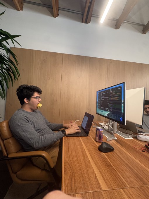 A man with glasses and a gray sweater sitting at a wooden desk, working on a laptop while looking at a large monitor displaying code, with an apple in his mouth and an energy drink on the desk.