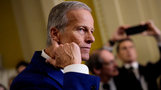 A distinguished older man with gray hair, wearing a dark suit, rests his hand on his chin with a thoughtful expression while someone in the background holds up a smartphone to take a photo. The contemplative pose visually reinforces the post's question about whether the crypto Clarity Act will pass before Friday's recess.
