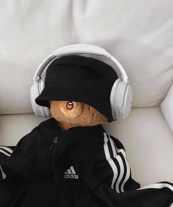 A brown teddy bear dressed in an Adidas black bucket hat, white Sony headphones, and a matching black Adidas track jacket sits on a light-colored sofa.