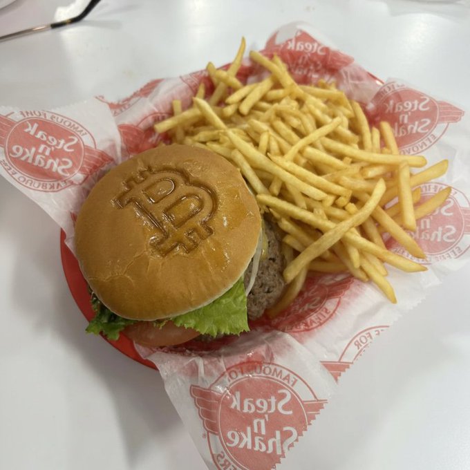 The post suggests that buying steak 'n shake is now a luxury purchase; the image shows a "Steak 'n Shake" branded burger and fries in a basket. The "Steak 'n Shake" name is visible in red lettering on the wrapper, along with "LAKELAND, FLA" and the interlocking "SnS" logo branded into the bun.