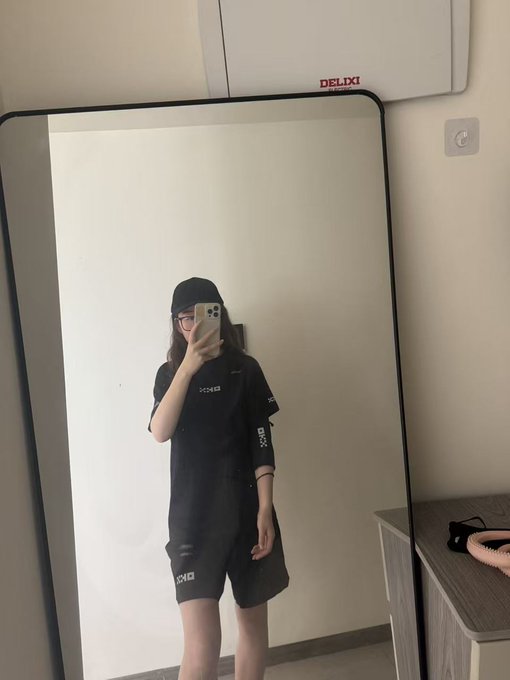 Mirror selfie of a person wearing glasses, a black beanie, and a black graphic t-shirt and shorts, holding an iPhone in front of a mirror with a DELIXI electrical box visible on the wall.