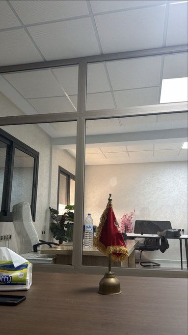 An office view through a glass partition, showing a desk with a water bottle, plants, and a chair, with a small red flag on a brass bell in the foreground.