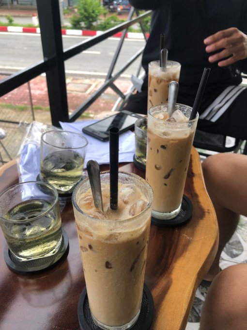 Multiple glasses of iced coffee and water with black straws on a wooden table at an outdoor café overlooking a street.