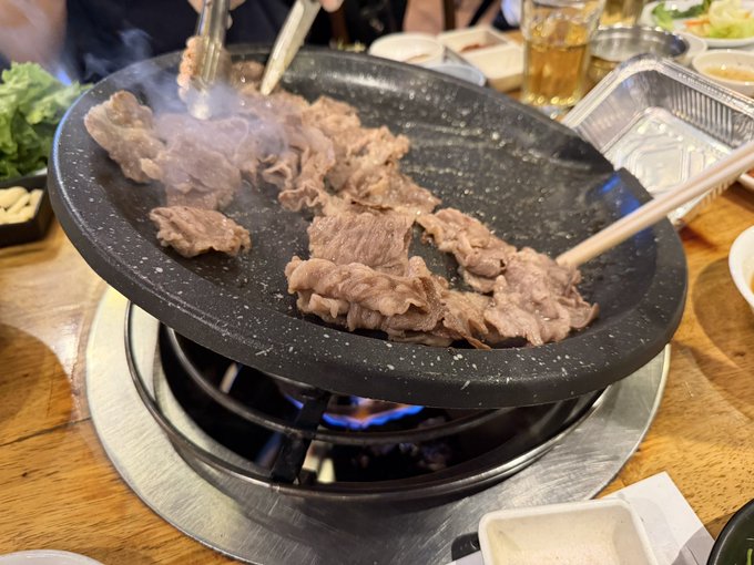 Thinly sliced beef cooking on a black stone tabletop grill over an open flame, with chopsticks handling the meat and side dishes visible around the grill.