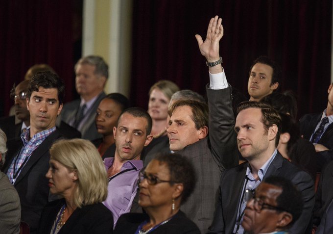 A diverse audience sits in an auditorium, with one man in a gray suit in the center raising his hand as if to ask a question, while Steve Carell is visible on the left side of the frame.