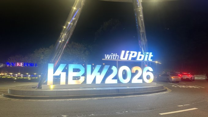 A nighttime photograph of an illuminated "KBW 2026 with Upbit" sign at the entrance to Korea Blockchain Week, with cars passing by.